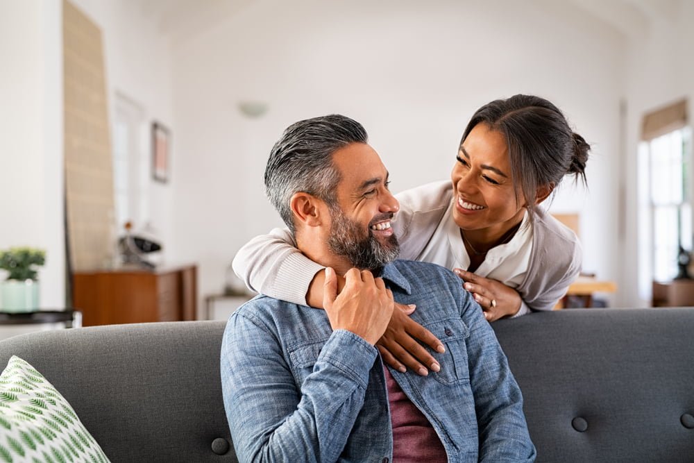 A couple shares a tender moment at home, with the woman leaning over the back of the couch to embrace the man from behind. Both are smiling warmly, looking at each other with affection. The scene is cozy and relaxed, set in a well-lit living room, reflecting a sense of comfort and connection - Dentist Georgetown TX A couple shares a tender moment at home, with the woman leaning over the back of the couch to embrace the man from behind. Both are smiling warmly, looking at each other with affection. The scene is cozy and relaxed, set in a well-lit living room, reflecting a sense of comfort and connection - Dentist Georgetown TX