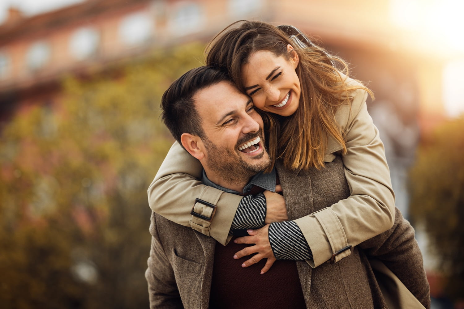 A couple joyfully embraces outdoors in the sunlight. The woman, smiling widely, wraps her arms around the man’s shoulders from behind as they both laugh - Dentist Georgetown TX A couple joyfully embraces outdoors in the sunlight. The woman, smiling widely, wraps her arms around the man’s shoulders from behind as they both laugh - Dentist Georgetown TX